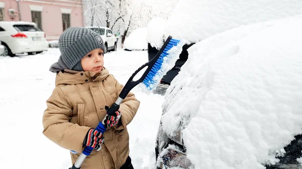 Zbliżenie strzał szczęśliwy chłopiec w płaszcz i szary kapelusz oczyścić biały samochód po śniegu upadku z pędzlem — Zdjęcie stockowe