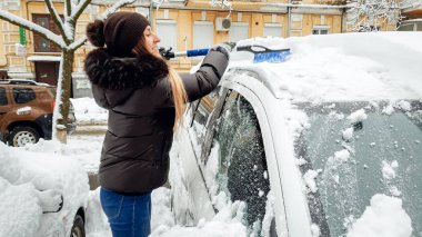 Siyah ceket, kot pantolon ve braun şapka kar yağışı sonrası mavi fırça ile karla kaplı araba temizlemek için çalışırken güzel gülümseyen kız yakın görüntü .