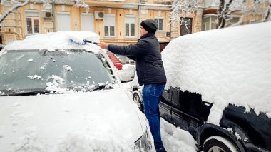 Sarı eve karşı fırça ile kar fırtınası sonra karla kaplı araba temizlemek için çalışırken ceket ve şapka adam tam uzunlukta fotoğraf