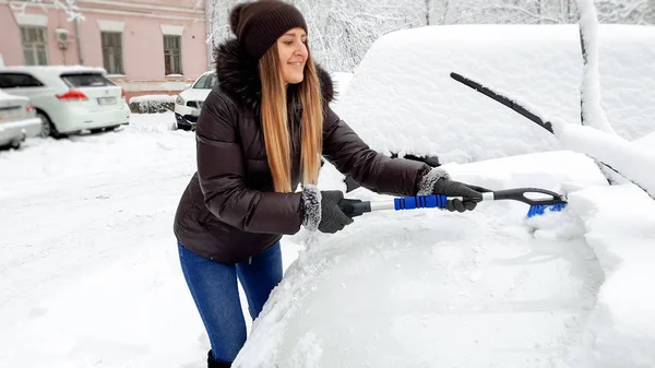 Retrato de la feliz señora rubia sonriente con abrigo marrón y sombrero tratando de limpiar el coche cubierto de nieve por el cepillo después de una ventisca —  Fotos de Stock