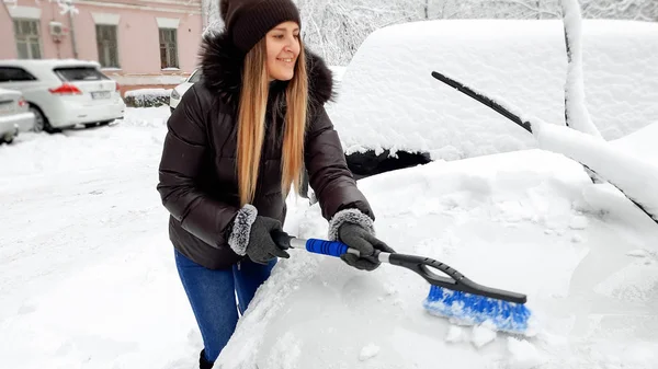 Foto di giovane donna sorridente in cappotto marrone e cappello che cerca di ripulire l'auto coperta di neve con la spazzola dopo una bufera di neve — Foto Stock