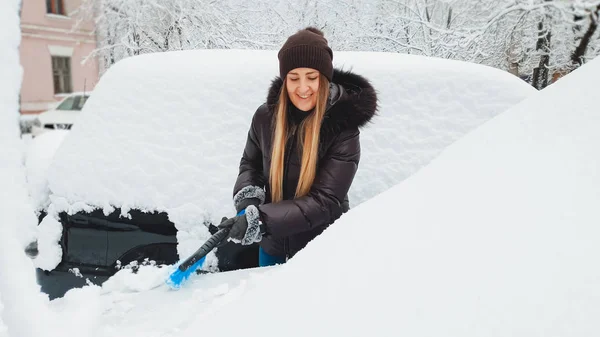 Закрыть изображение молодой красивой женщины в коричневом пальто и шляпе, очищающей машину и стеклоочистители от снега щеткой после снежной бури — стоковое фото