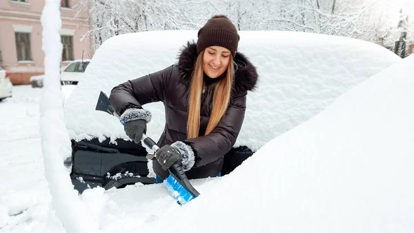 Fotografie usmívající se krásná dívka v hnědém kabátě a klobouku vyčistí auto a stírače čelního skla ze sněhu kartáčem po sněhové bouři — Stock fotografie