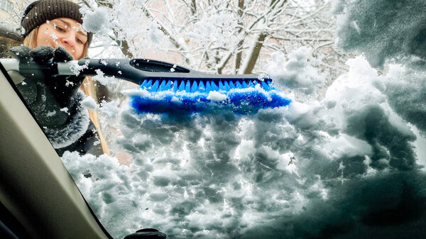 Close up photo inside the snow covered car. Beautiful young woman in brown hat trying to clean up the windshield and wipers of her auto after blizzard