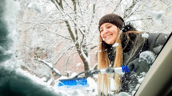 Zbliżenie strzał wewnątrz śniegu pokryte samochód. Uśmiechnięta dziewczyna stara się oczyścić przednią szybę i wycieraczki jej auto po burzy śnieżnej z pędzlem — Zdjęcie stockowe