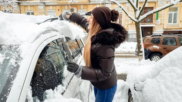 Zbliżenie wizerunek młodego preaty uśmiechnięta blond dama w czarnym płaszczu niebieskie dżinsy i kapelusz próbuje oczyścić śnieg pokryte samochodu przez niebieski pędzel po śniegu upadku . — Zdjęcie stockowe