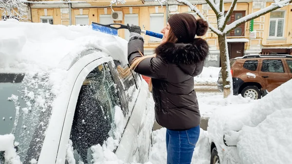 Primo piano colpo di preatra sorridente signora bionda in nero cappotto blu jeans e cappello cercando di pulire auto innevata da spazzola blu dopo la caduta della neve  . — Foto Stock
