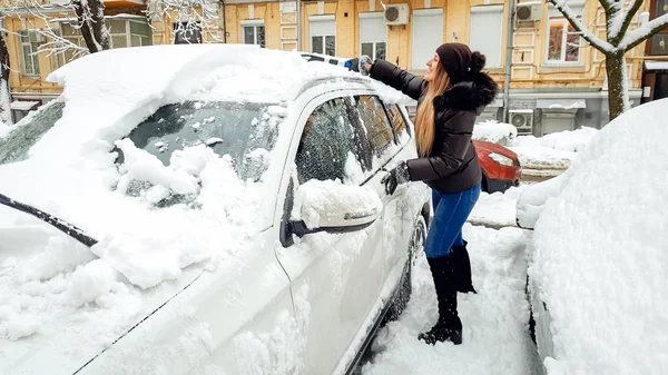 Shot of leende ung kvinna i päls, jeans och Braun hatt försöker städa upp snötäckt bil med pensel efter snöfall . — Stockfoto