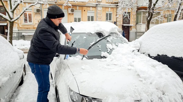 Shot of man i svart jacka försöker städa upp bilen från snö med borste mot huset — Stockfoto