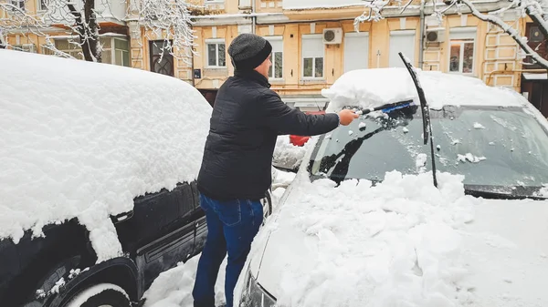 Pełna długość obrazu człowieka w płaszczu i kapeluszu próbuje oczyścić śnieg pokryte samochód po burzy śnieżnej z pędzlem przeciwko domu — Zdjęcie stockowe