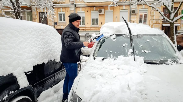 Выстрел красивого человека в пальто и шляпе, пытающегося очистить заснеженный автомобиль после метели с черной кистью — стоковое фото