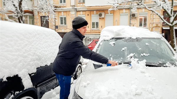 Foto de un hombre guapo con abrigo y sombrero tratando de limpiar el coche cubierto de nieve después de ventisca con cepillo — Foto de Stock