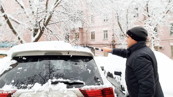 Крупным планом фотография молодого красивого мужчины в черном пальто и шляпе, пытающегося очистить белую машину от снега после метели кистью — стоковое фото