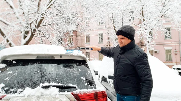 Närbild skott av ung stilig man i svart päls och hatt försöker städa upp snötäckt bil efter snöstorm med borste — Stockfoto