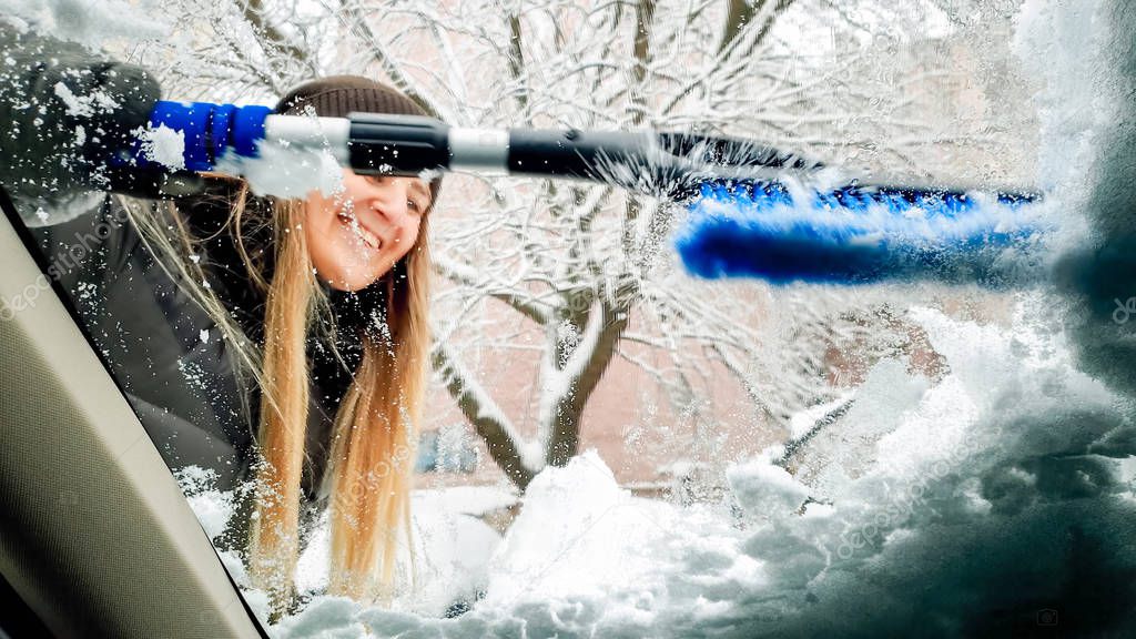 Closeup portrait inside the snow covered car. Beautiful young smiling woman in hat trying to clean up the windshield and wipers of her auto after blizzard with blue brush