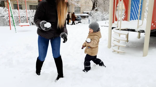 Image de jeune mère heureuse playind en boules de neige avec son mignon fils en veste et chapeau sur l'aire de jeux dans le parc — Photo