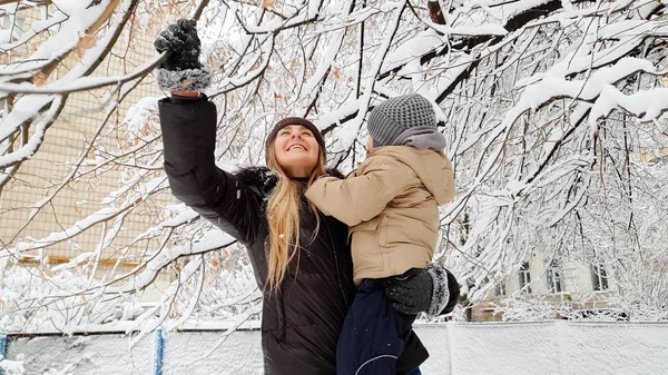 Bliska portret kobiety z jej cute syna w marynarce i kapeluszu gry z pokrytym śniegiem drzewa na placu zabaw w parku — Zdjęcie stockowe