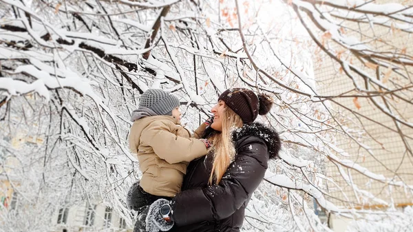 Close-up beeld van gelukkig glimlachende jonge moeder in bruine vacht en muts met haar mooie zoon in beige jasje spelen met besneeuwde boom op de speelplaats in het Park — Stockfoto