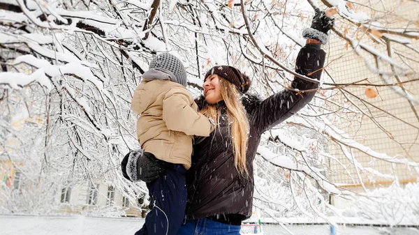 Primer plano de feliz sonriente joven madre en abrigo marrón y sombrero con su bonito hijo en chaqueta jugando con el árbol cubierto de nieve en el patio de recreo en el Parque — Foto de Stock
