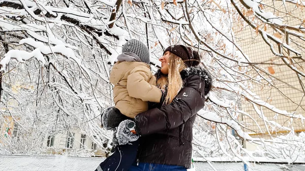 Bliska obraz szczęśliwy uśmiechnięta młoda matka w płaszcz i kapelusz z jej ładny syn w marynarce gry z pokrytym śniegiem drzewa na placu zabaw w parku — Zdjęcie stockowe