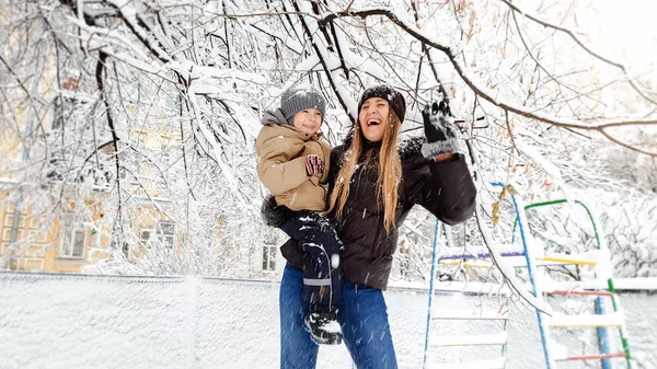 Retrato de cerca de una joven y sonriente madre con abrigo y sombrero con su hermoso hijo con chaqueta jugando con un árbol cubierto de nieve en el patio del parque — Foto de Stock