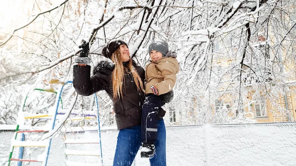 Portrait rapproché d'une jeune mère souriante avec son joli fils en veste et chapeau jouant avec un arbre recouvert de neige sur l'aire de jeux du parc — Photo