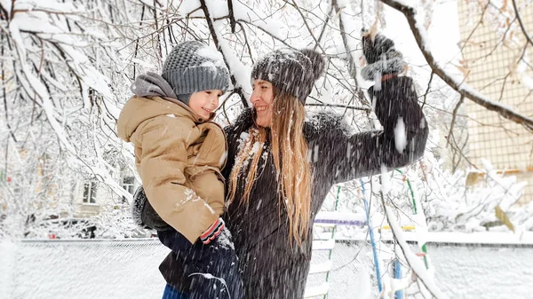Zblízka se rozveselila mladá matka s hezkým synem ve svěrací kazajce a klobouku, které si pohrává s sněhem pokrytou stromem na hřišti v parku. — Stock fotografie