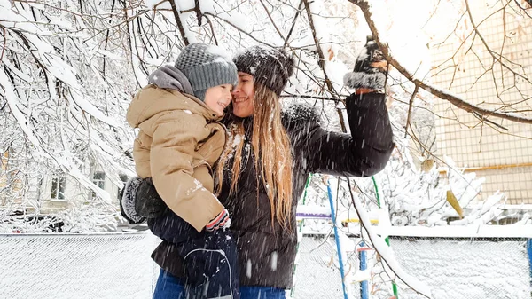 Foto de cerca de la alegre madre joven con su lindo hijo en chaqueta y sombrero jugando con el árbol cubierto de nieve en el patio de recreo en el Parque — Foto de Stock