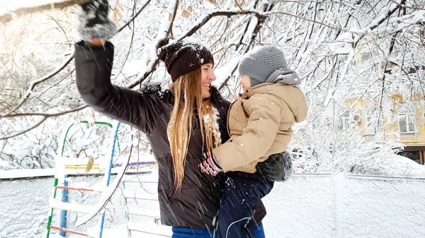 Imagen de cerca de la joven rubia con su lindo hijo en chaqueta y sombrero jugando con el árbol cubierto de nieve en el patio en el Parque — Foto de Stock