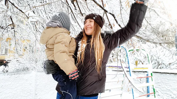Zbliżenie strzał blond kobieta z jej cute syna w marynarce i kapelusz gry z pokrytym śniegiem drzewa na placu zabaw w parku — Zdjęcie stockowe
