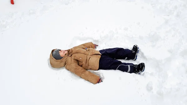 Foto de niño lindo en abrigo, divertirse a jugar en la nieve después de ventisca. Juega en bolas de nieve en el parque infantil en el parque. Como tumbarse en la nieve — Foto de Stock