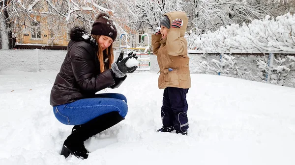 De cerca foto de feliz sonriente joven madre en abrigo de broun y sombrero con su hijo bonito en chaqueta beige divertirse a jugar las bolas de nieve con nieve en el patio de recreo en el Parque — Foto de Stock