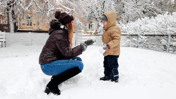 Immagine primo piano di allegra giovane madre sorridente in cappotto broun e cappello con il suo bel figlio in giacca beige divertirsi a giocare le palle di neve con la neve sul parco giochi nel parco — Foto Stock