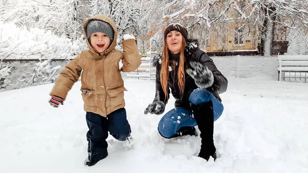 Bliska strzał wesoły uśmiechnięta młoda matka w płaszcz i kapelusz z jej ładny syn w beżowej kurtki baw się bawić piłki śnieżne z śniegu na placu zabaw w parku — Zdjęcie stockowe