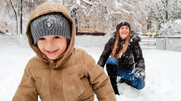 微笑的母亲在外套与她可爱的儿子在米色夹克有乐趣玩雪与雪在公园的游乐场的特写图像 — 图库照片