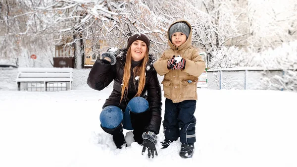 Zbliżenie zdjęcie uśmiechnięta młoda matka w płaszcz i kapelusz z jej cute syna w beżowy kurtka baw się bawić piłki śniegu z śniegu na placu zabaw w parku — Zdjęcie stockowe