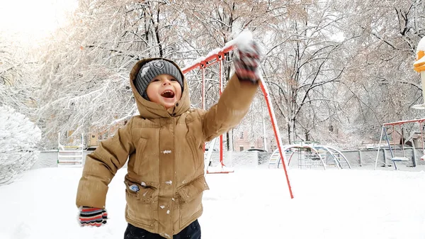 Gros plan d'un garçon mignon et souriant en manteau, amusez-vous à jouer dans la neige en hiver après les chutes de neige. Jouer dans les boules de neige sur l'aire de jeux au parc — Photo