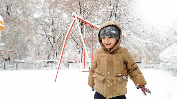 Bliska strzał uśmiechnięty szczęśliwy cute Boy w beżowy płaszcz i szary kapelusz, baw się bawić w śniegu w zimie po śniegu. Graj w kule śnieżne na placu zabaw w parku — Zdjęcie stockowe