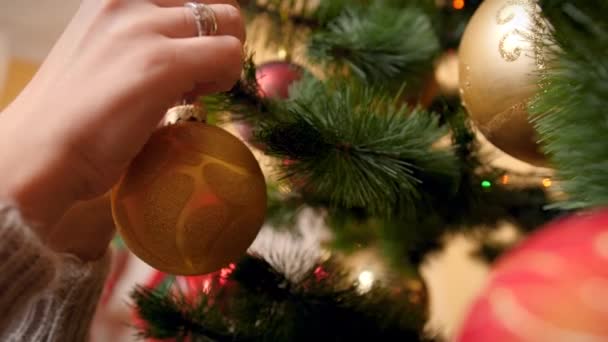 Primer plano 4k video de mujer joven colgando y girando oro brillante adornado bauble en el árbol de Navidad. Personas preparándose para las celebraciones de invierno y días festivos . — Vídeo de stock