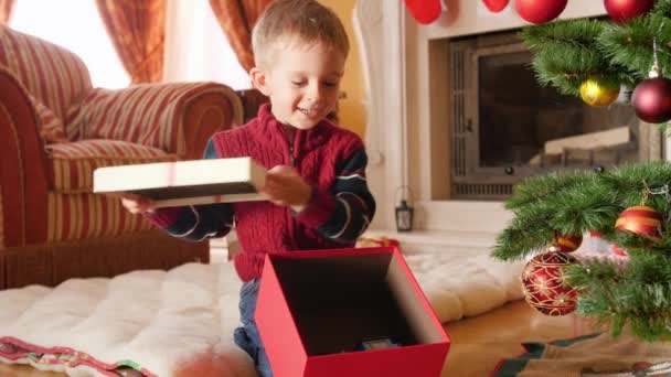 4k Video von fröhlich lächelndem kleinen Jungen, der den Deckel aus der Weihnachtsgeschenkbox nimmt und die Spielzeugeisenbahn herausnimmt, die ihm der Weihnachtsmann geschenkt hat. Kind erhält Geschenke und Geschenke in den Winterferien und — Stockvideo