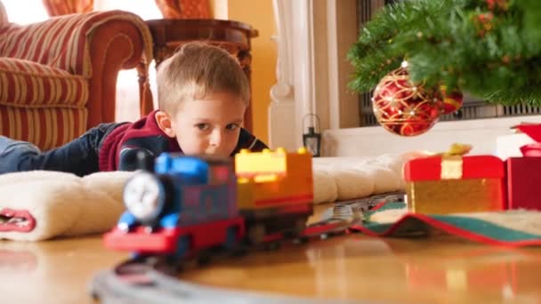4k video de niño mintiendo en el suelo bajo el árbol de Navidad y mirando el tren de juguete cabalgando en el ferrocarril. Niño recibiendo regalos y regalos de Santa Claus en las vacaciones de invierno y celebraciones — Vídeos de Stock