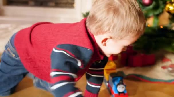 Vidéo 4k de petit garçon souriant qui s'amuse et joue avec le train jouet et le chemin de fer sous l'arbre de Noël dans le salon. Enfant recevant des cadeaux et cadeaux du Père Noël pendant les vacances d'hiver et — Video