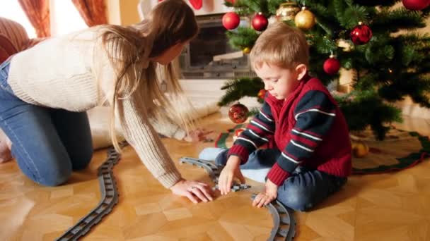 4k metraje de feliz madre sonriente ayudando a su hijo a construir la vía férrea de ladrillos bajo el árbol de Christams. Niño recibiendo regalos y regalos de Santa Claus en las vacaciones de invierno y celebraciones — Vídeos de Stock