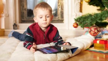 4k video of little boy lying on blanket under Christmas tree and playing video games on digital tablet computer. Child having fun on winter holidays and celebrations.