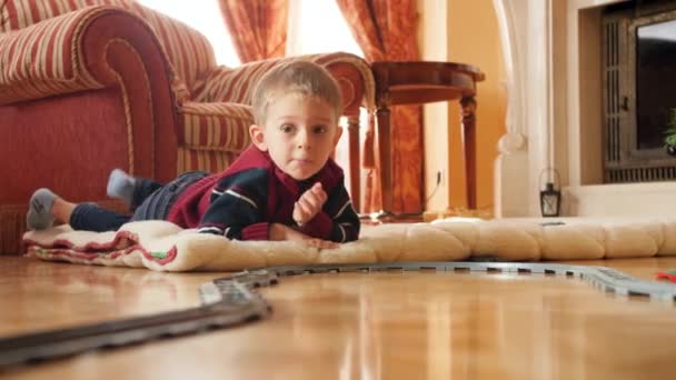 Vidéo 4k de petit garçon souriant jouant avec le chemin de fer jouet et train sur le sol dans le salon le matin de Noël. Enfant recevant des cadeaux et des cadeaux du Père Noël pendant les vacances et les célébrations d'hiver — Video