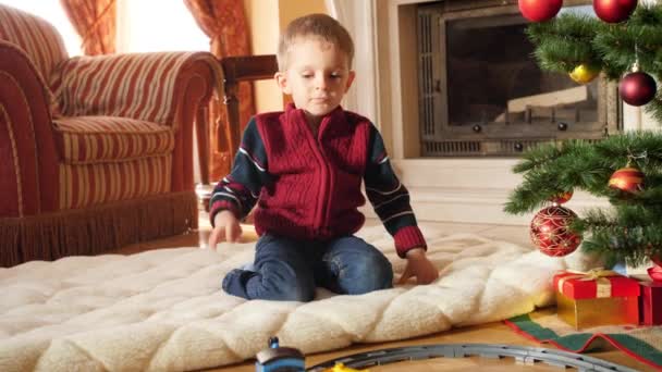 Imágenes de 4k del niño pequeño mirando el tren de juguete cabalgando en el ferrocarril circular en la sala de estar. Niño recibiendo regalos y regalos de Santa Claus en las vacaciones de invierno y celebraciones — Vídeo de stock