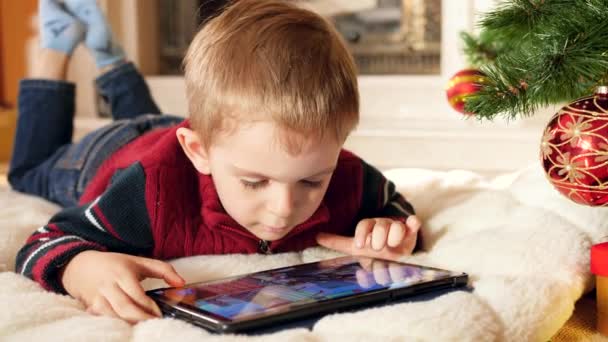 Primer plano 4k video de un niño sonriente jugando juegos en una tableta digital mientras yacía bajo el árbol de Christams en casa. Niño divirtiéndose en vacaciones de invierno y celebraciones . — Vídeo de stock