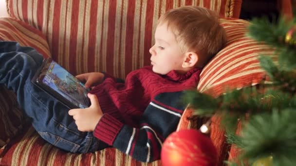 4k Nahaufnahme Video von kleinen Jungen, die im Sessel im Wohnzimmer liegen und Videospiele auf einem digitalen Tablet-Computer spielen. Kinder haben Spaß beim Winterurlaub und Feiern. — Stockvideo