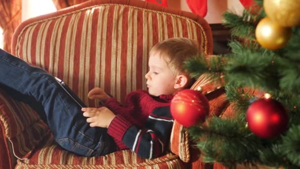 Images 4k de petit garçon allongé dans un fauteuil le matin de Noël et jouant à des jeux sur tablette numérique 