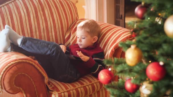Vídeo 4k de menino deitado no sofá na sala de estar e usando o computador tablet. Criança se divertindo nas férias de inverno e celebrações . — Vídeo de Stock